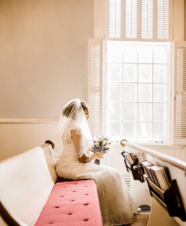 A bride sits in a church pew, holding her bouquet near a bright window. She is wearing a white dress and veil.