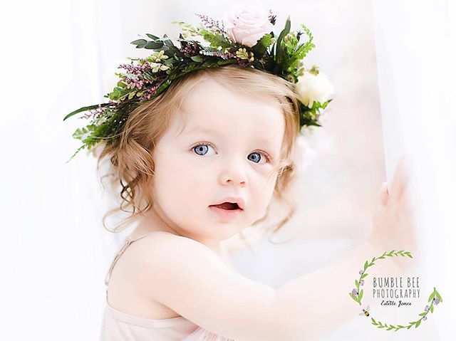 A sweet toddler with blue eyes wears a flower crown in a soft, dreamy portrait.