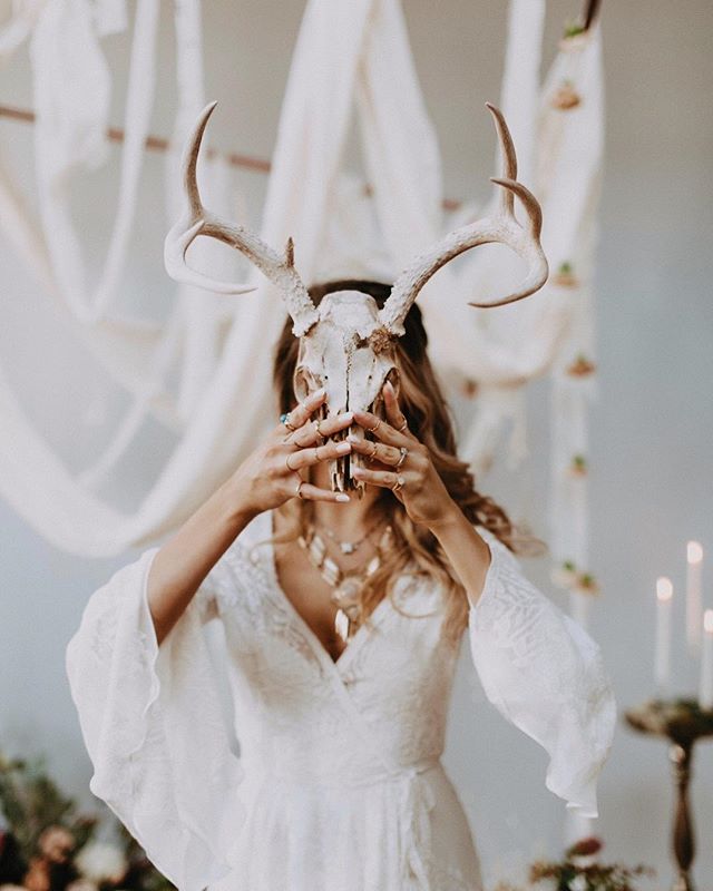 A woman in a white dress holds a deer skull with antlers, adorned with jewelry for a bohemian style wedding.