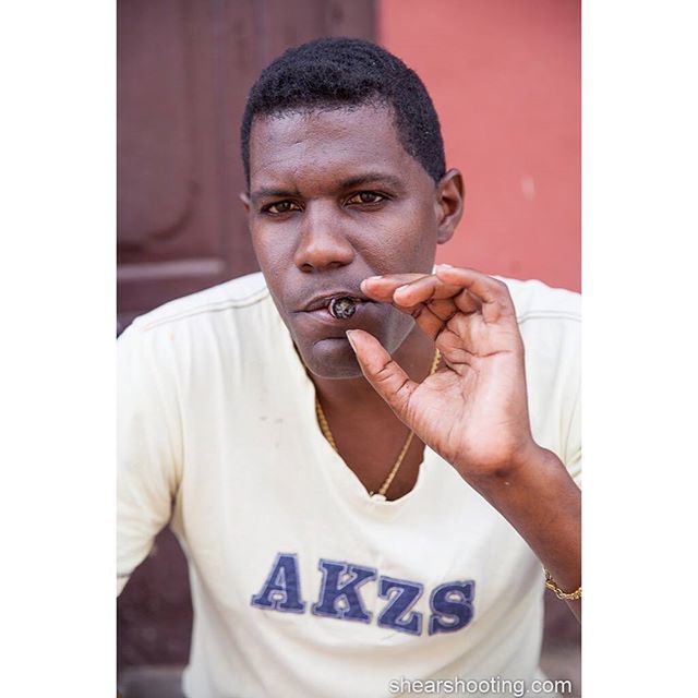 A man is smoking a cigar outside while wearing a white shirt and gold chain.