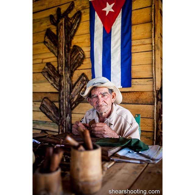 An elderly Cuban man sits at a table rolling cigars in a rustic workshop beneath a Cuban flag.