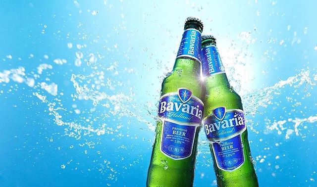 Two Bavaria beer bottles clinking together with a splash of water, set against a bright blue background, perfect for a refreshing ad.