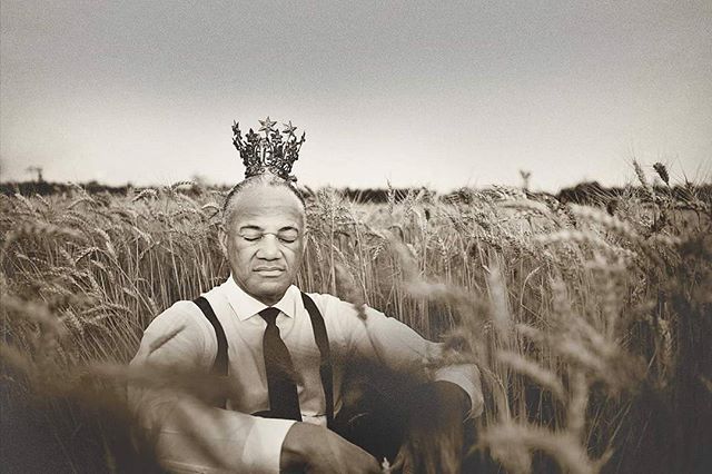 Man with crown sits in wheat field wearing shirt and tie in a vintage style.