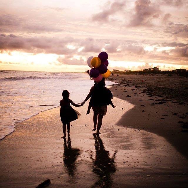 A mother and child walk hand-in-hand along the beach at sunset, carrying colorful balloons.