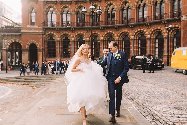 A bride and groom walk together with the wedding party outside a brick building on their wedding day.
