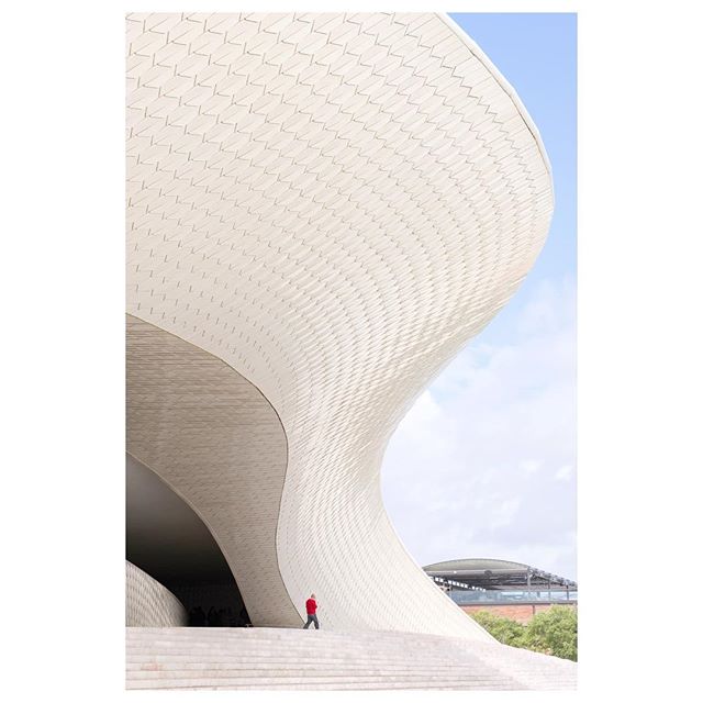 A person walks up the stairs of a modern, curved building with a textured white facade against a bright sky.