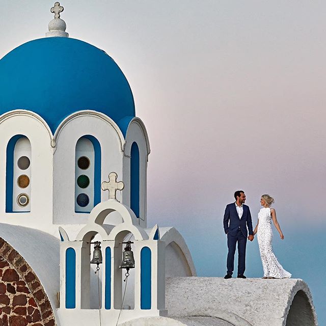 A bride and groom stand on a rooftop with a blue-domed church in Santorini, Greece.
