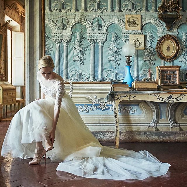 A bride in a white wedding dress puts on her shoe in a vintage-style room.