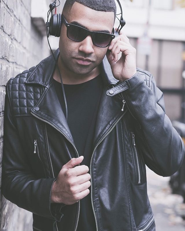A stylish man in sunglasses and a leather jacket leans against a brick wall while listening to music with headphones.