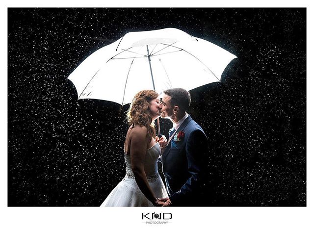 A bride and groom share a kiss under an umbrella in the rain, a romantic moment captured in a wedding photograph.