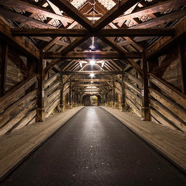 The inside of a wooden covered bridge with an asphalt road, featuring a detailed wooden beam ceiling.