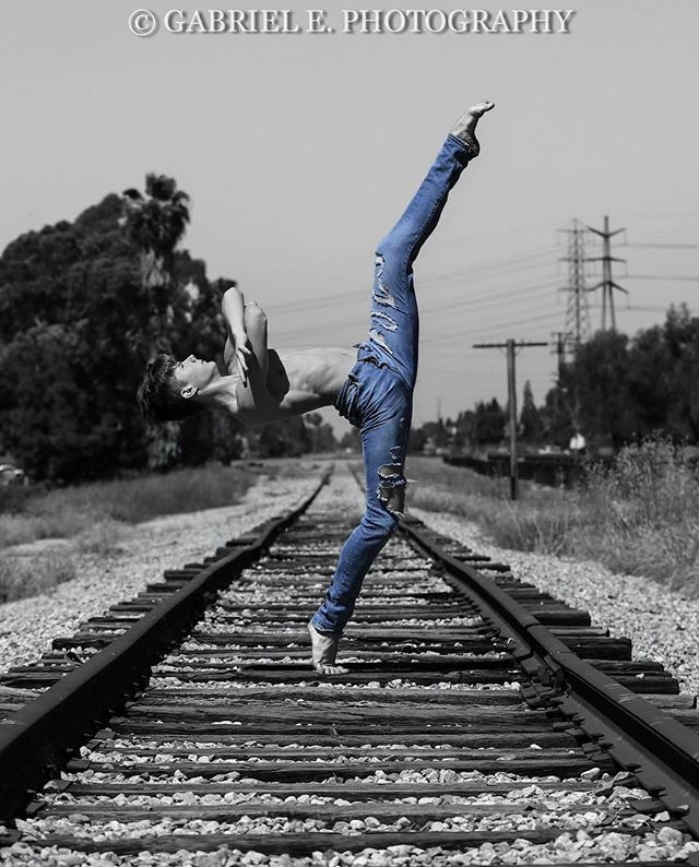 A man in jeans performs a dynamic dance move on railroad tracks in a captivating black and white image with selective color.