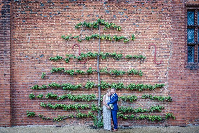 A happy bride and groom pose in front of a beautiful brick wall and tree espalier.