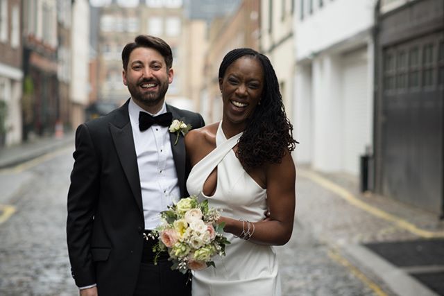 A smiling bride and groom pose on a city street with a bouquet, celebrating their wedding day.
