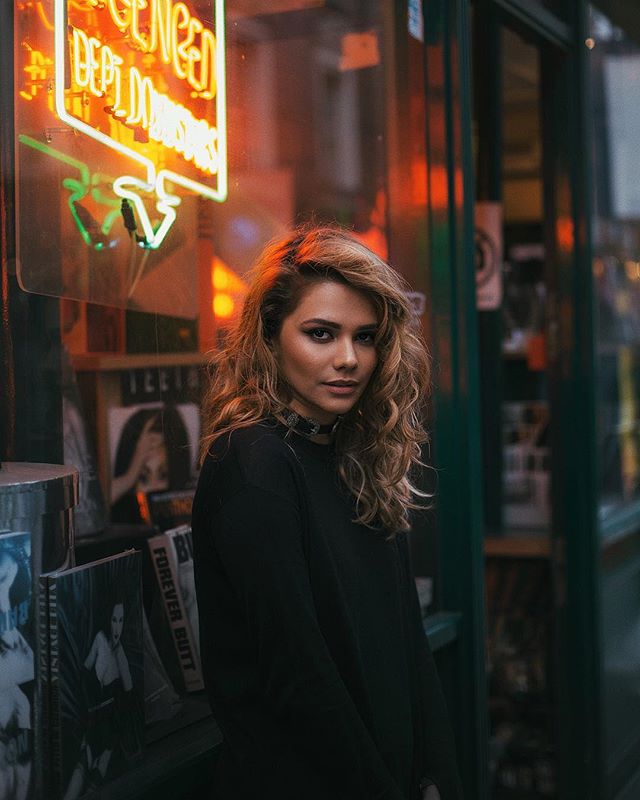 A young woman with curly hair stands by a store window with a neon sign, creating a moody portrait.