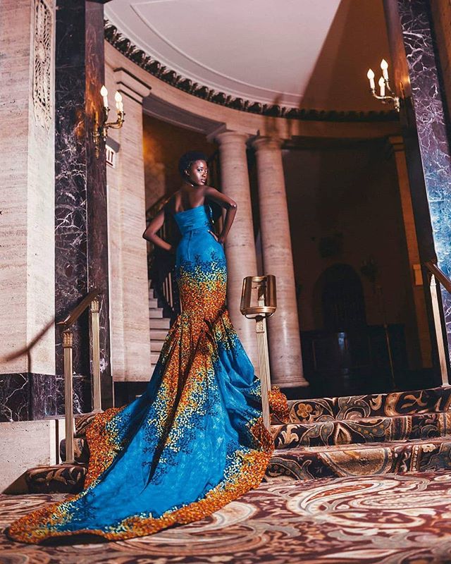 A woman in a blue and orange gown stands on stairs in an elegant building.