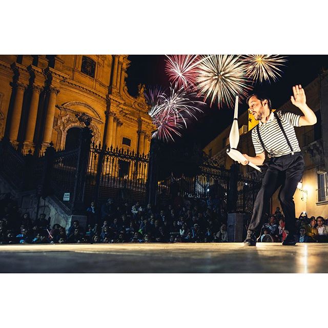 A juggler performs at night with fireworks in the background, entertaining a large audience.