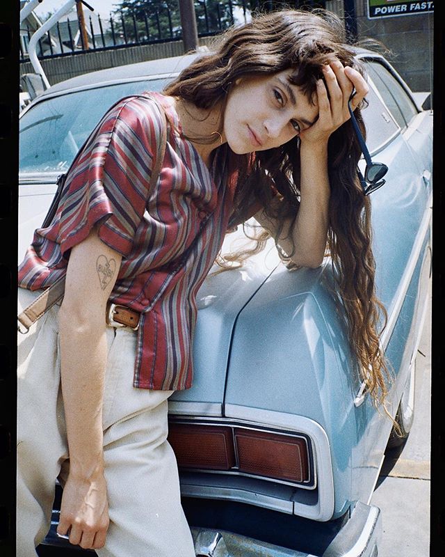 A young woman poses leaning on a vintage car in an urban setting during daytime.