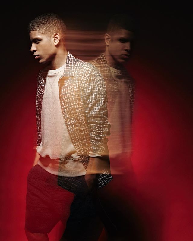A stylish man poses in a studio with a red background in a fashion editorial shot.