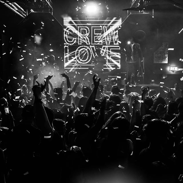 A lively crowd celebrates at a nightclub with confetti and a stage performance in black and white.