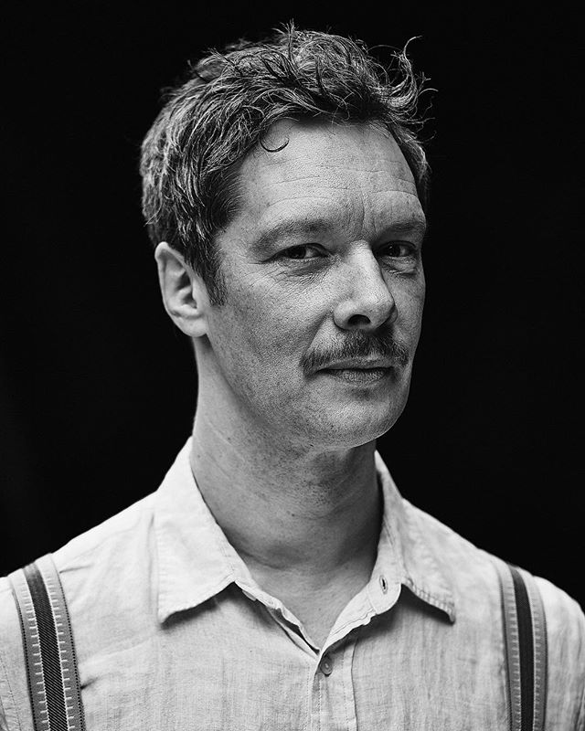 A black and white portrait of a man with a mustache, wearing suspenders against a dark background.