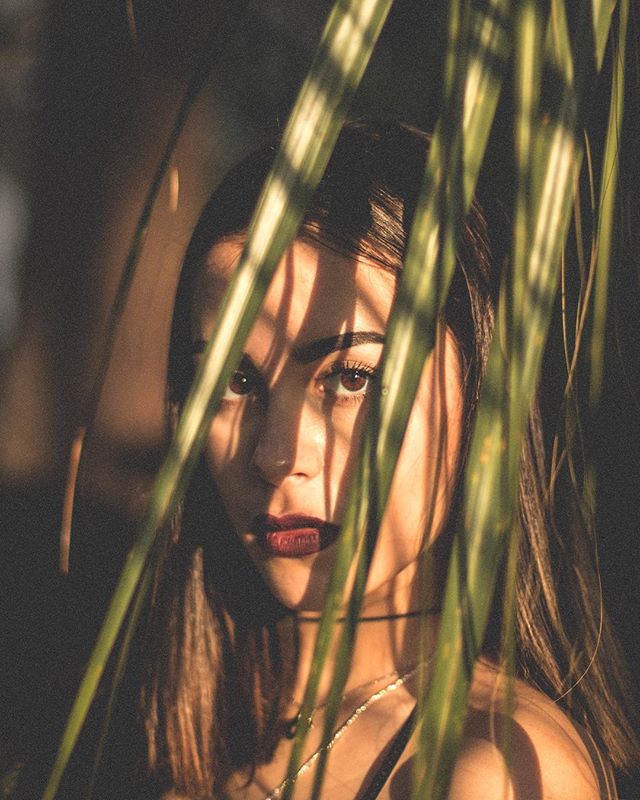 A woman with dark lips poses behind green leaves in soft natural light, creating a mysterious and moody portrait.