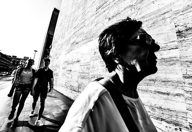 A black and white street photo featuring people walking alongside a tall building, bathed in sunlight.