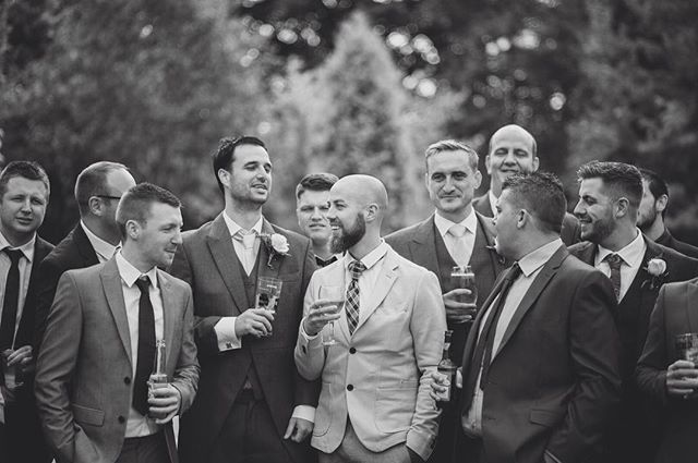 A black and white image captures a large group of groomsmen celebrating outdoors at a wedding.