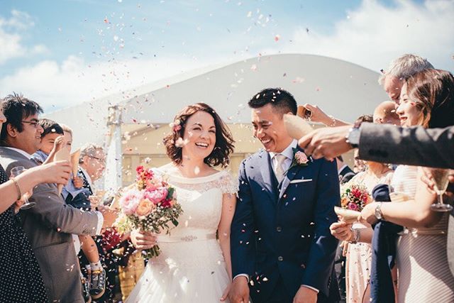 A smiling bride and groom are showered with confetti by their guests at their outdoor wedding reception.