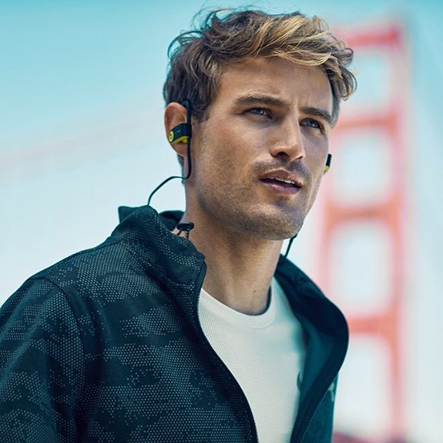 A fit man with earphones poses in an urban outdoor setting. He is wearing fitness clothing.