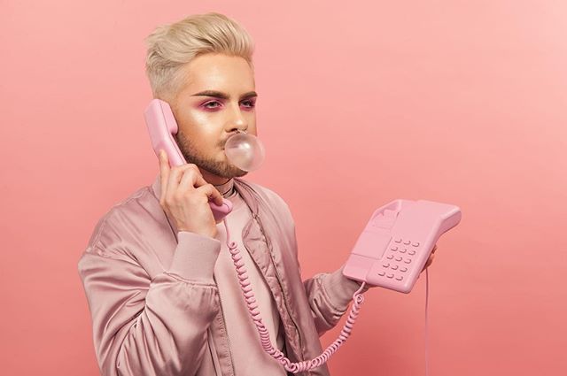 A man with bubble gum poses with a pink telephone in a studio setting with a pink background.