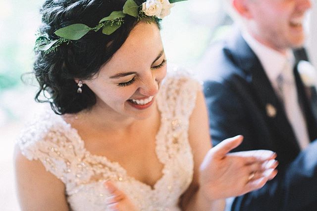 A joyful bride smiles during her wedding reception, clapping her hands with a man blurred in the background.