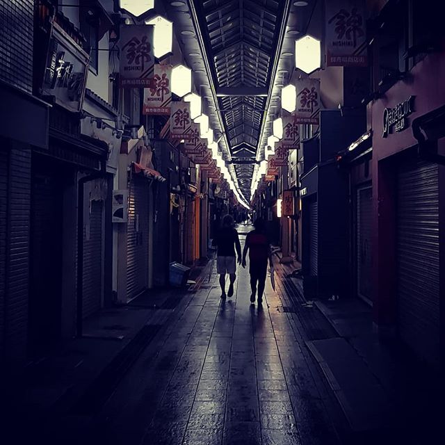 Two people walk through a dark, covered alleyway illuminated by hanging lanterns at night.