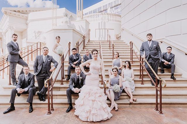 A wedding party poses on stairs outside, featuring the bride, groom, bridesmaids, and groomsmen in formal attire.