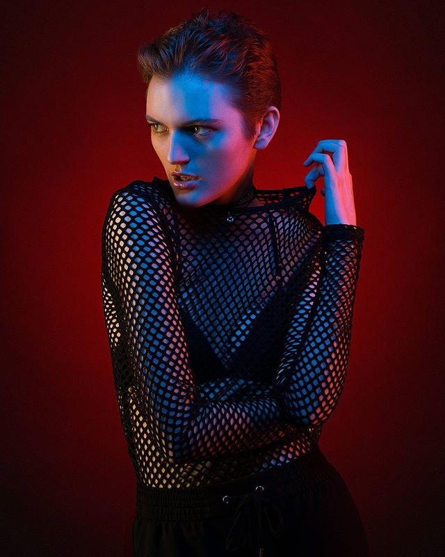 A woman with short hair poses in a fishnet top against a vibrant red background.