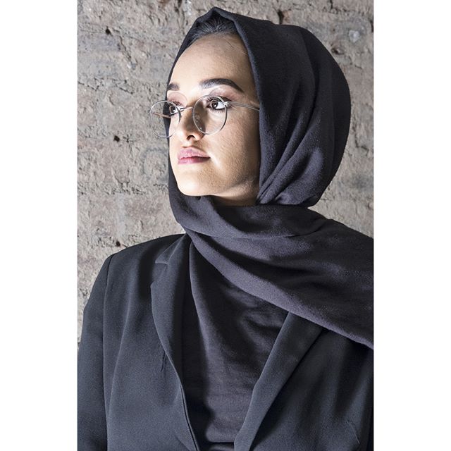 A woman wearing a hijab and glasses poses against a textured wall in a studio shot.