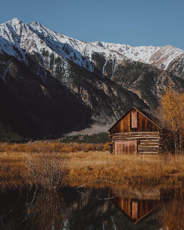 A rustic cabin sits by a serene lake, framed by snow-capped mountains in an autumnal landscape.