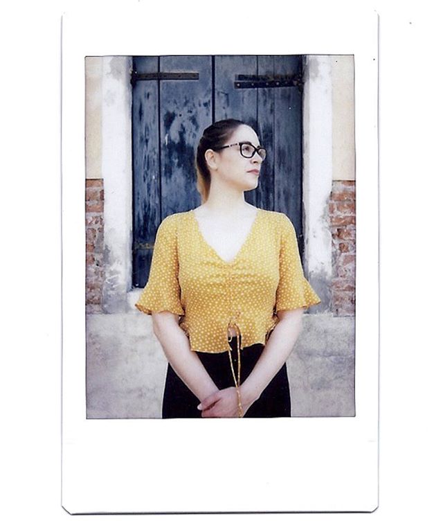 A woman wearing glasses and a yellow polka dot top poses in front of a blue door.