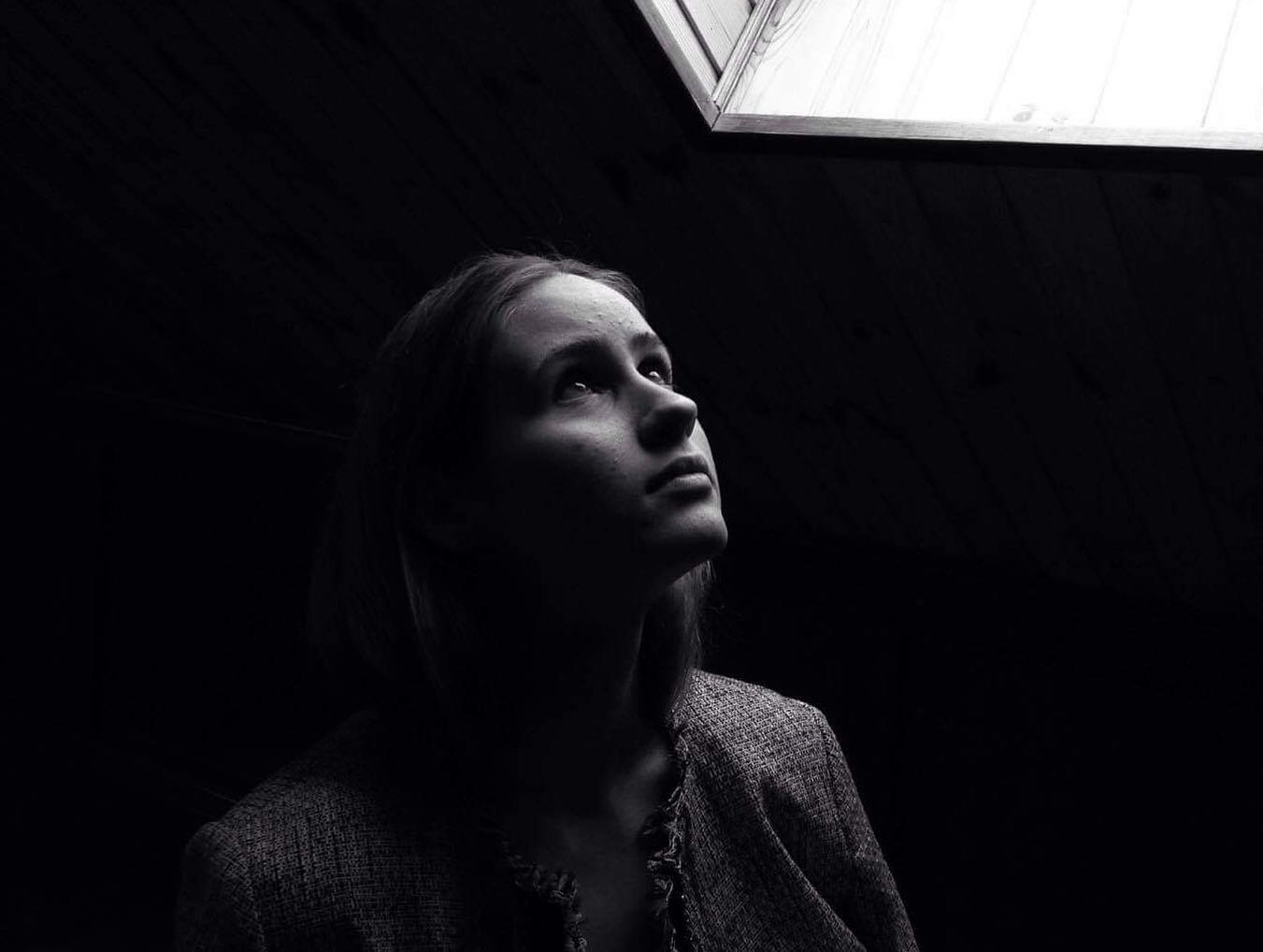 A young woman looks up toward a light source in a dark room, creating a high-contrast black and white portrait.