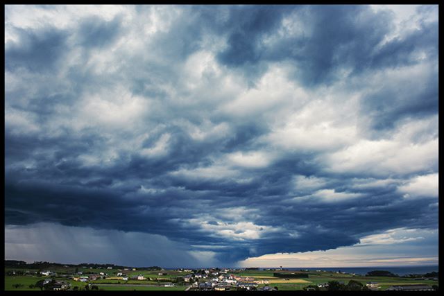 A dramatic storm cloudscape hovers above a small town nestled in a green countryside landscape.