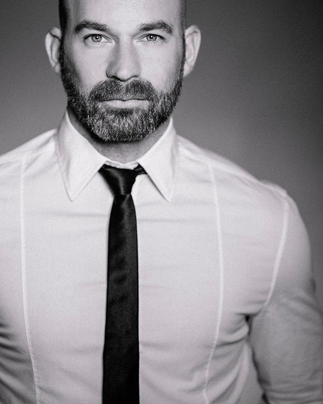 A confident man with a beard is wearing a shirt and tie for a professional business portrait in black and white.