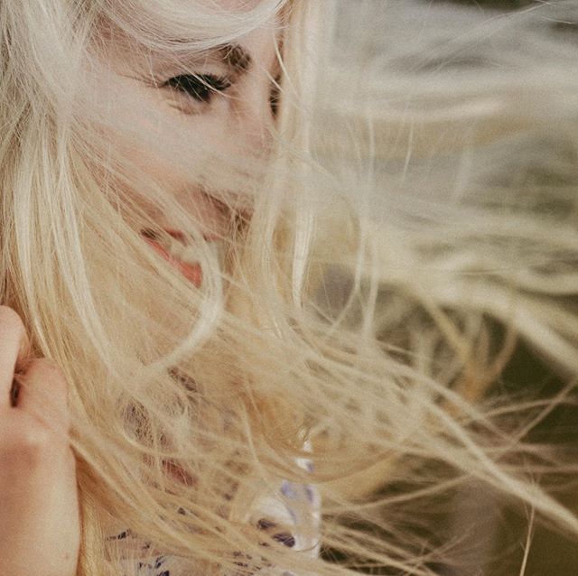 A blonde woman with windswept hair smiles softly in a sunlit field, creating a dreamy, serene atmosphere.