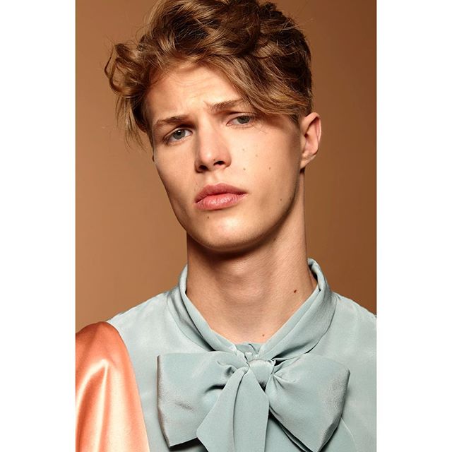 A young man poses in a studio wearing a light blue bow tie shirt, shot in soft lighting.