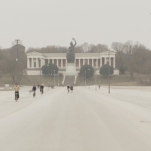 People cycle near the Monopteros monument and building in a park in Munich, Germany on an overcast day.
