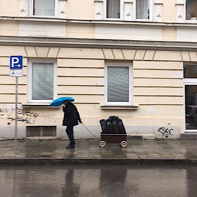 A person pulls a cart full of tires in the rain under a blue umbrella on a city street.