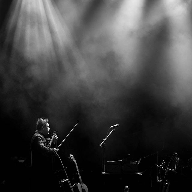 A musician performs with a violin on a dimly lit stage in black and white.