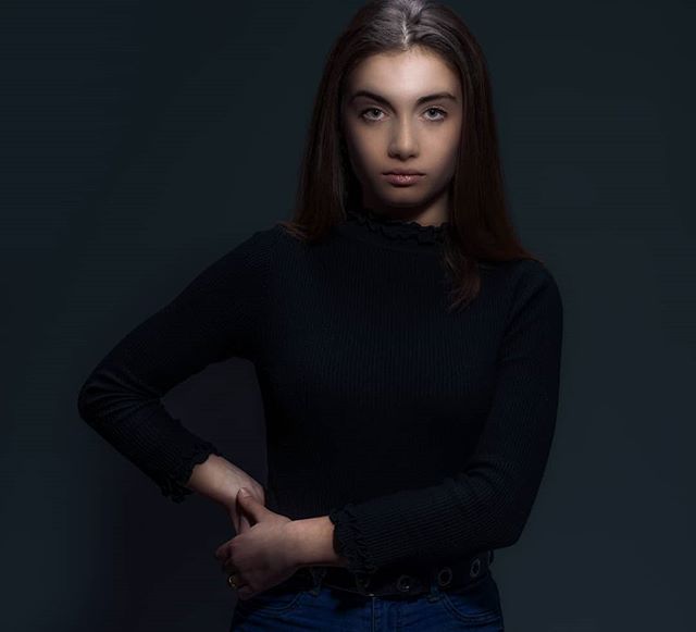 A young woman with long brunette hair poses confidently in a studio setting with soft, flattering light.