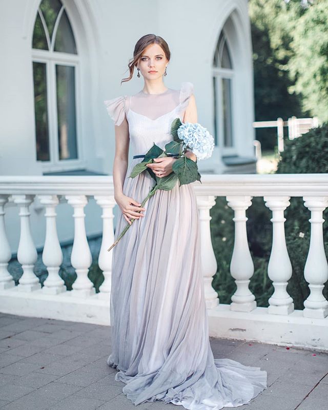 A woman in a grey dress holds a blue hydrangea on a balcony, exuding elegance.