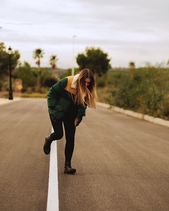 A woman balances on a white line in the road, wearing a green and tan jacket in a casual outdoor setting.