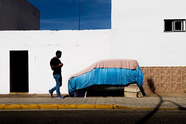 A man walks by a car covered with a blue tarp on a sidewalk in an urban setting on a bright day.